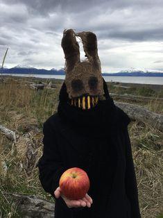 a person wearing a bunny mask holding an apple in front of the camera with mountains in the background