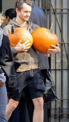 a man holding two pumpkins in his hands while walking down the street with other people behind him