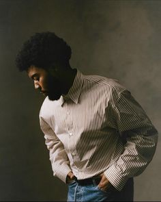 a man with his hands on his hips wearing a striped shirt and jeans, standing in front of a gray background