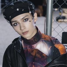 a woman with piercings on her head wearing a leather jacket and black hat, standing in front of a chain link fence