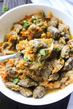 a white bowl filled with meat and vegetables on top of a wooden table next to a spoon