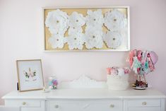 a white dresser topped with lots of flowers next to a wall mounted flower art piece