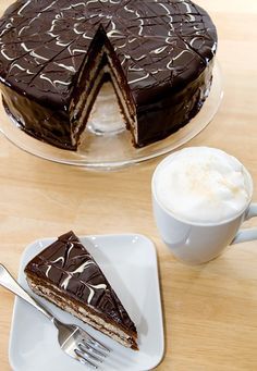 a chocolate cake on a plate with a slice missing from it and a cup of cappuccino in the background