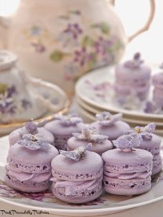 there are many purple macaroons on the plate and tea pot in the background