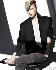 a man sitting on top of a stool next to a white wall wearing a black blazer