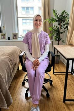 Female medical professional sitting at a desk in a bedroom, wearing the Easy STRETCH by Butter-Soft™ Cara Women's 4-Pocket Raglan Scrub Top and the Mila Women's 5-Pocket Jogger Scrub Pants Purple Scrubs, Mist Color