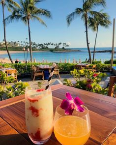 two drinks sitting on top of a wooden table next to the ocean and palm trees