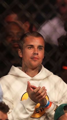 a man wearing a white hoodie sitting in front of a fence with his hands clasped