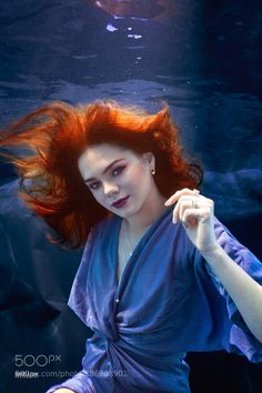 a woman with red hair sitting under the water in a blue dress looking at the camera