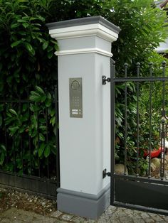 a white clock on the side of a black fence next to some bushes and trees