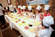 a group of children sitting at a table with pizzas on it and chefs in the background