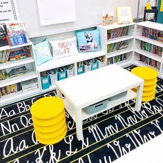 a room filled with lots of books and yellow stools next to a white table