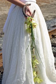 a woman in a white dress holding flowers and a bird on her arm while standing next to train tracks