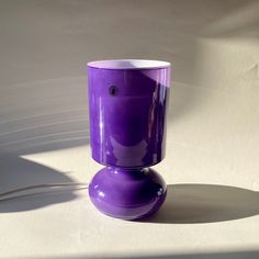 a purple vase sitting on top of a white table next to a light shadow from the wall