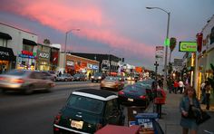 cars are driving down the street in front of shops and businesses at dusk with pink clouds