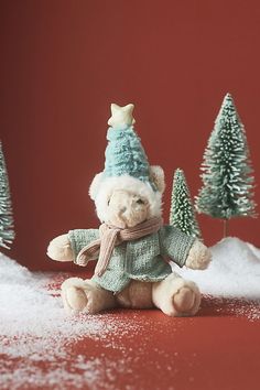 a teddy bear wearing a hat and scarf sitting in the snow next to small trees