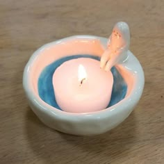 a small white candle in a bowl on a wooden table with a little figurine