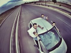 three men sitting on the hood of a silver sports car driving down a road with mountains in the background