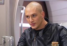 a bald man sitting at a table in front of a blender and glass bottle