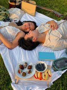 a man and woman laying on a blanket in the grass next to some fruit, chocolates and strawberries