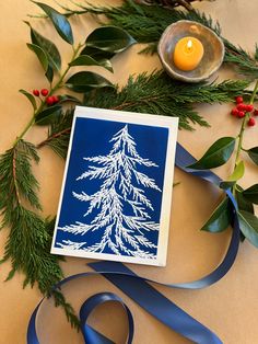 a card with a white pine tree on it next to blue ribbon and holly branches