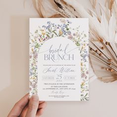 a person holding up a wedding card with flowers and feathers around it on top of a table