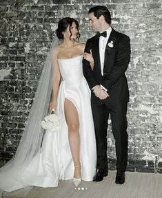 a bride and groom standing next to each other in front of a brick wall wearing tuxedos