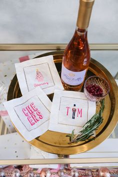 three embroidered napkins on a gold tray next to a bottle of wine and some ornaments