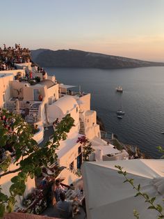 people sitting at tables on the edge of a cliff overlooking the ocean and boats in the water