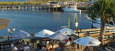 people are sitting at tables near the water with umbrellas and boats in the background