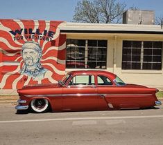 an old red car parked in front of a building with a mural on it's side