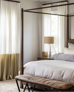 a bedroom with a four poster bed and white curtains on the window sill, along with a bench in front of it