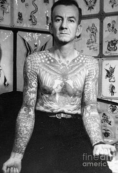 a man with tattoos on his chest standing in front of a wall full of pictures