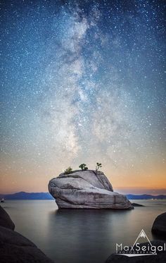 the night sky is filled with stars above a large rock in the middle of water