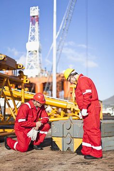 two men in red work suits and hardhats at a construction site with machinery behind them