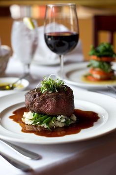 a white plate topped with meat covered in sauce and garnish next to a glass of wine