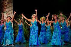 a group of women in blue dresses are dancing on stage with their arms raised up