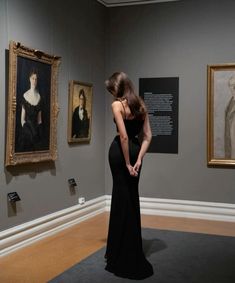 a woman in a black dress looking at some paintings on the wall and holding her hands behind her back