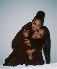 two girls hugging each other on a bed together, one is wearing a brown sweater and the other has long black hair