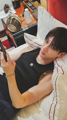 a young man laying in bed holding a cell phone and looking at the screen with his right hand