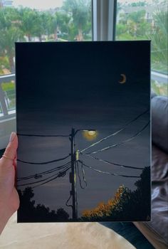 a person holding up a painting in front of a window with the moon above it