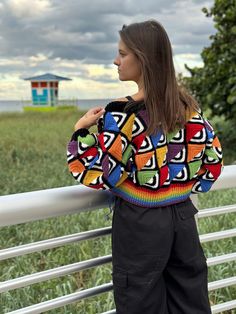 a woman standing next to a white fence wearing a colorful sweater and black pants with her hands on her hips