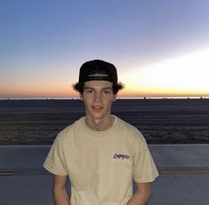 a young man standing in front of the ocean at sunset wearing a cap and t - shirt