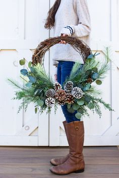 a woman holding a wreath with pine cones and evergreens