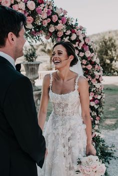 a bride and groom smile as they stand under an arch decorated with pink flowers at their wedding