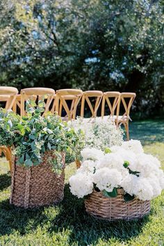 white flowers in wicker baskets on the grass near wooden chairs and tables for an outdoor ceremony