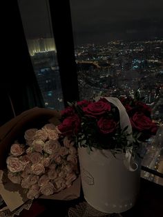 roses in a white bucket on top of a table next to a window overlooking a city at night