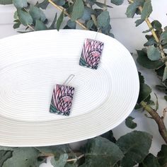 two pink and green earrings sitting on top of a white plate next to some leaves