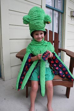 a young boy in a green costume sitting on a wooden bench wearing a hat and scarf