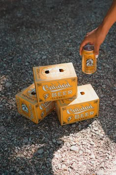 a person reaching for three cans of beer on the ground next to rocks and gravel